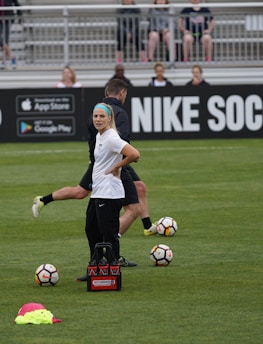 A person in sports attire stands on a soccer field, surrounded by several soccer balls and a carrier with bottles. The field is bordered by advertisements for Nike Soccer and app stores. Several people sit in bleachers in the background.