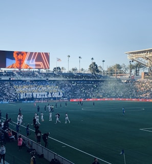A large sports stadium filled with spectators. The field is occupied by soccer players, some walking and others scattered across the pitch. A massive screen displays a person, and flags fly on top of the stadium roof. The audience is dense, and the atmosphere seems lively with a visible banner showing 'BLUE WHITE & GOLD'.